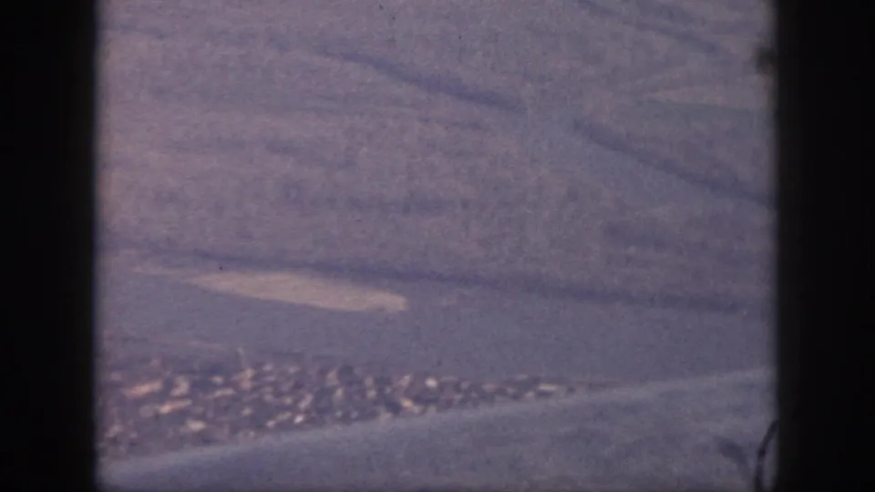 Aerial 8mm view over Oahu, with a pale shoreline, small developed area, water, and dark aircraft-window or gate-edge framing.