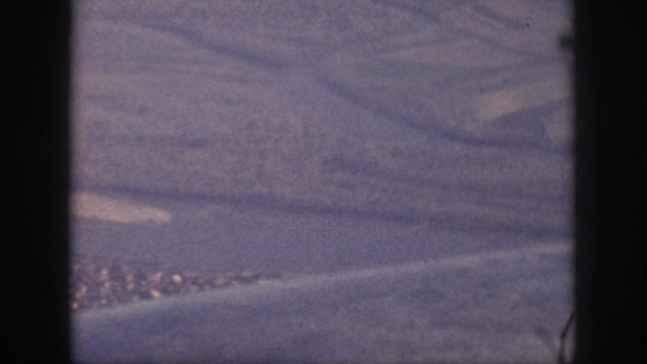 1959 Oahu airplane view with a blurred stretch of coastline, water, and open terrain framed by dark 8mm gate edges.