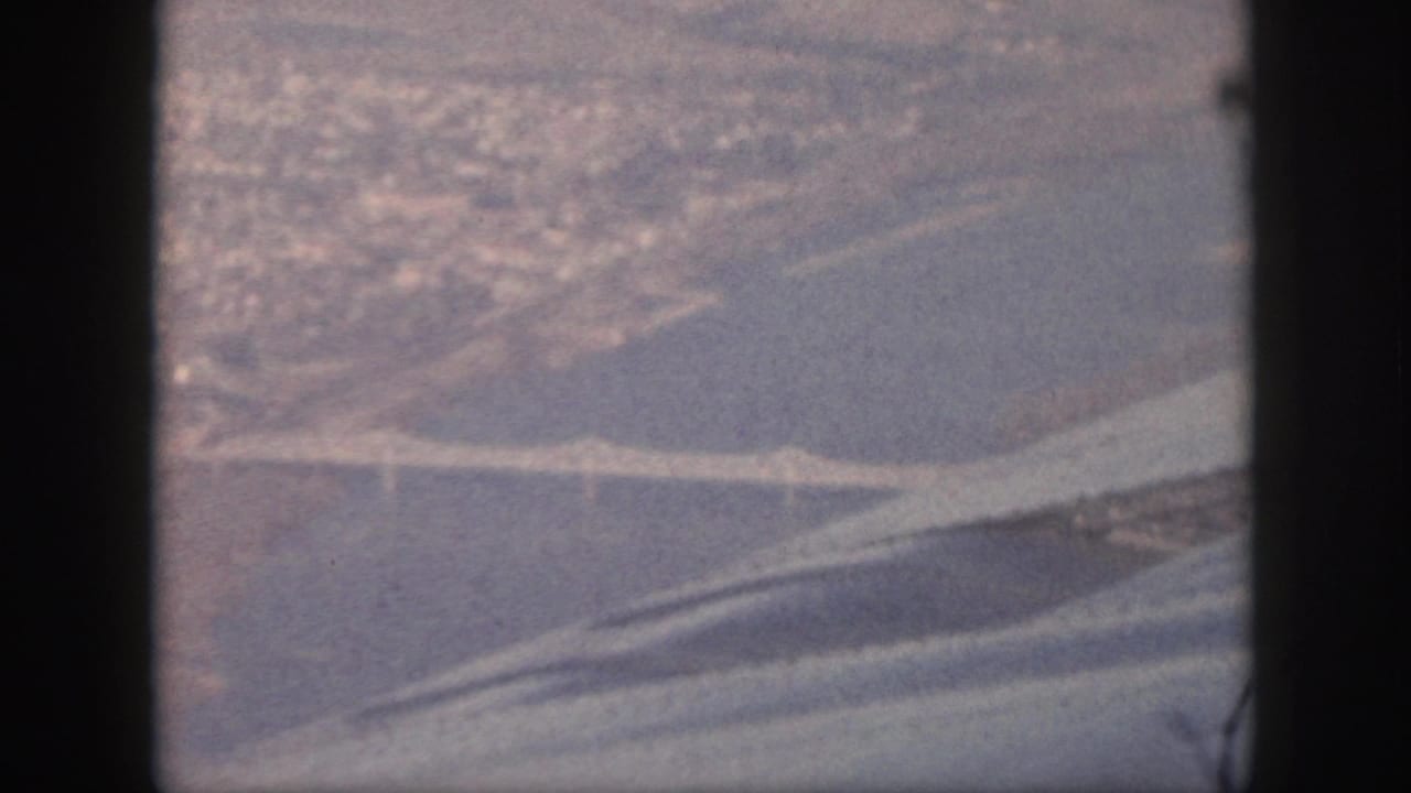 1959 Oahu 8mm aerial view with a bridge over a broad waterway and distant shoreline seen through an airplane window.
