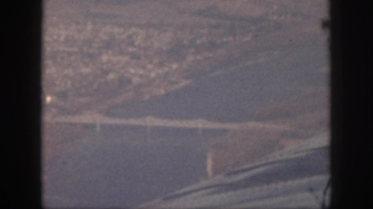 1959 Oahu aerial frame showing a bridge crossing water, with distant development softened by haze and film grain.