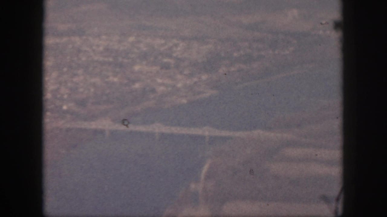 Final 1959 Oahu aerial frame with the water crossing, shoreline, and distant buildings visible through grain and haze.