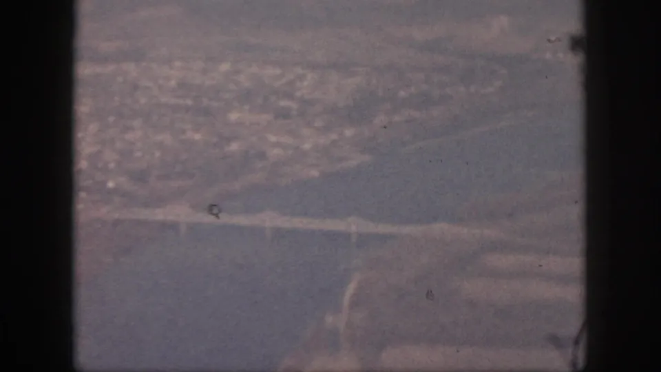 Final 1959 Oahu aerial frame with the water crossing, shoreline, and distant buildings visible through grain and haze.