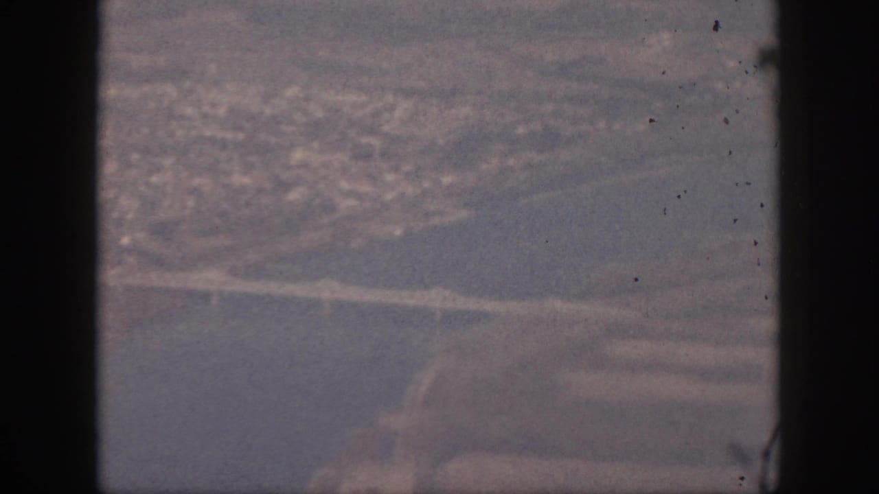 1959 Oahu aerial home-movie frame seen through an airplane window, with a river or inlet, bridge, roads, and hazy city area below.