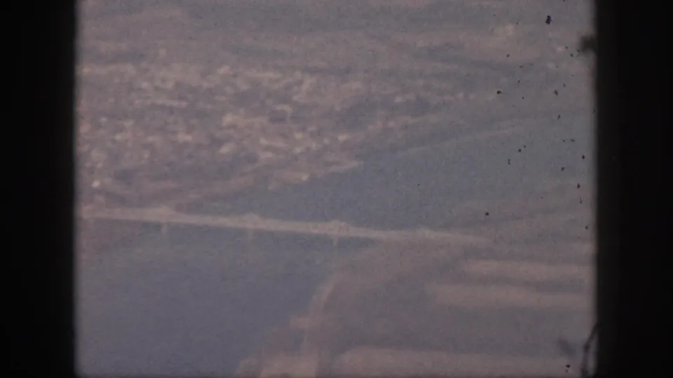 1959 Oahu aerial home-movie frame seen through an airplane window, with a river or inlet, bridge, roads, and hazy city area below.