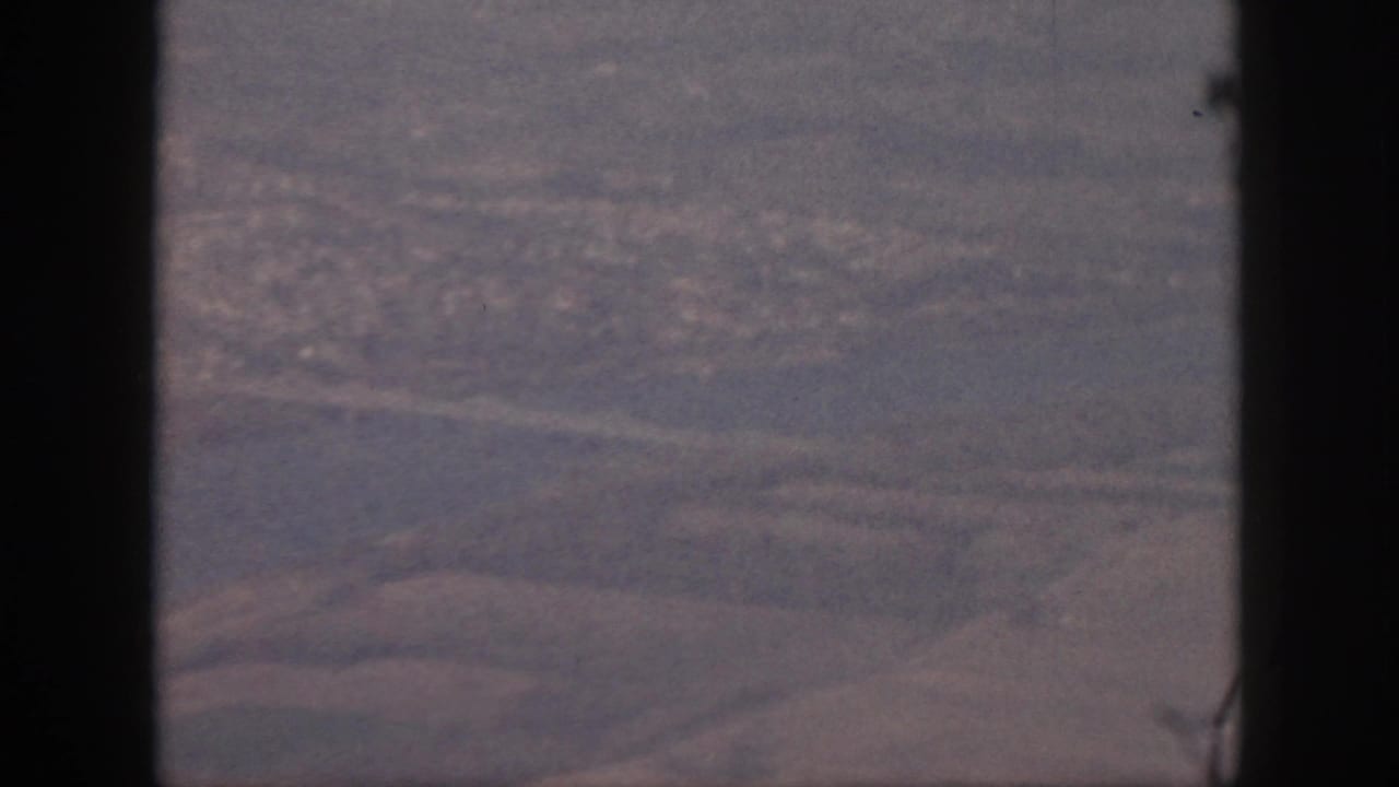 1959 Oahu aerial frame with the plane window frame prominent, showing muted water, roads, shoreline, and a distant built-up area below.