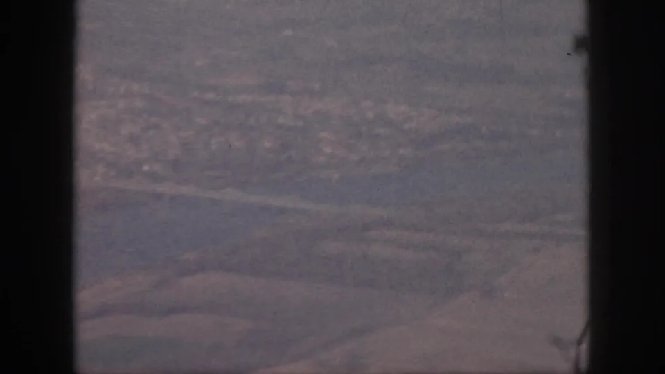 1959 Oahu aerial frame with the plane window frame prominent, showing muted water, roads, shoreline, and a distant built-up area below.