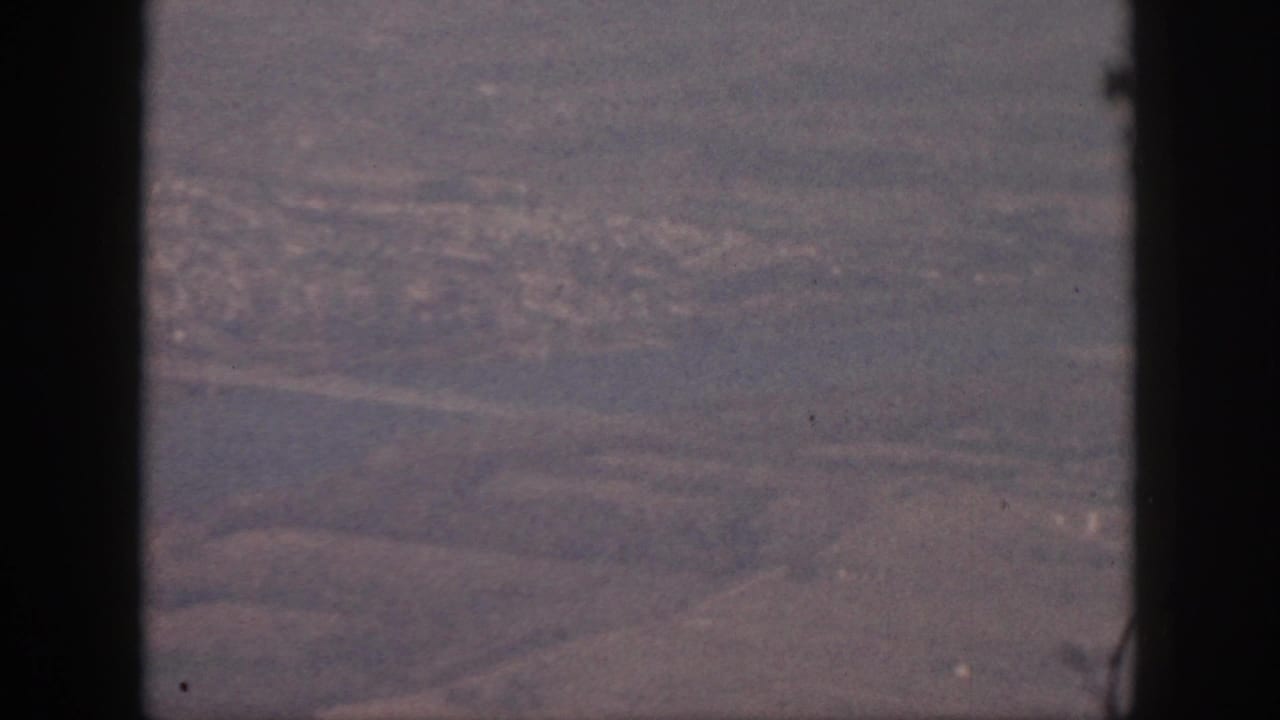 1959 Oahu home-movie aerial view through airplane-window edges, with a waterway, pale roads, shoreline, and distant urban texture.