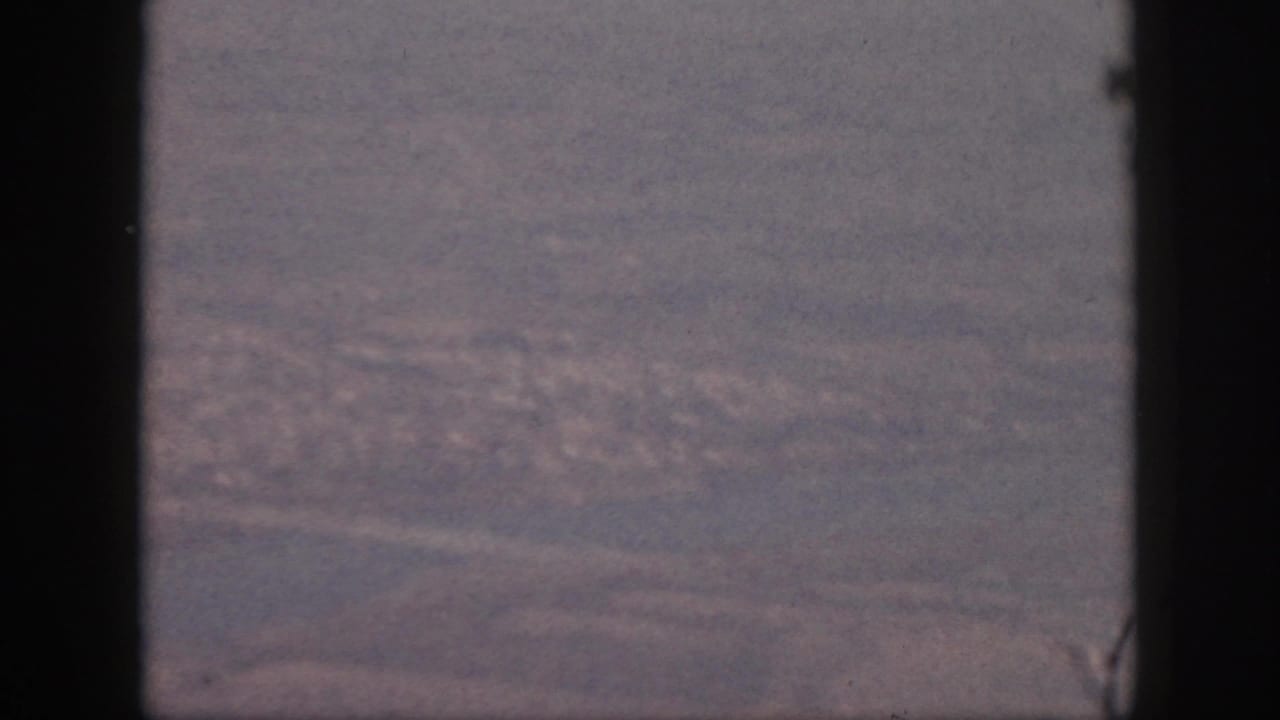 1959 Oahu aerial view from a plane window, showing water, shoreline, pale roads, and a distant cityscape in faded 8mm color.