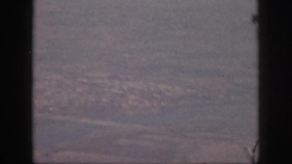 1959 Oahu aerial view from a plane window, showing water, shoreline, pale roads, and a distant cityscape in faded 8mm color.