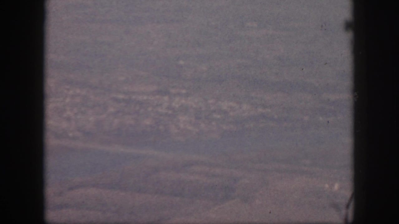 1959 Oahu home-movie frame showing a soft, purple-tinted aerial view of land or water through dark window-like bars.