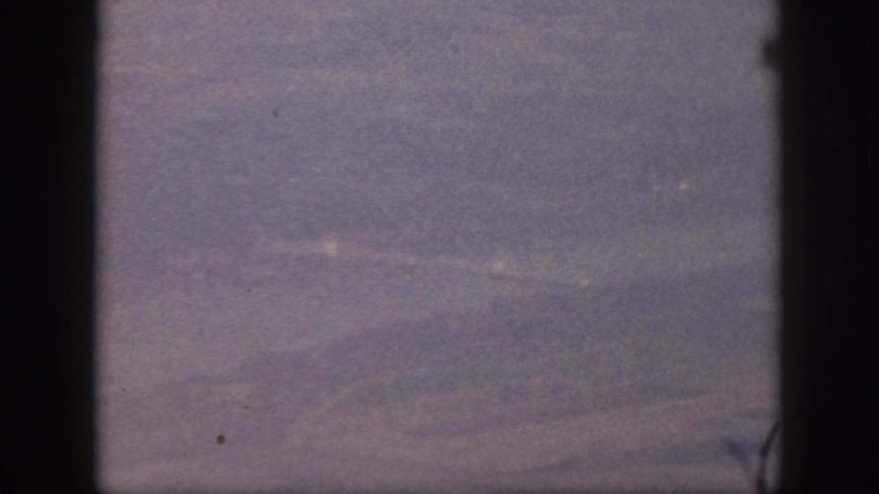 Purple-faded 8mm frame of an airplane-window view over indistinct Oahu terrain, with heavy grain and dark gate or window edges.