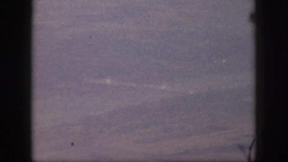 Purple-faded 8mm frame of an airplane-window view over indistinct Oahu terrain, with heavy grain and dark gate or window edges.