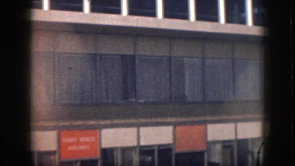 1959 Oahu home-movie frame of a modern building exterior with upper window band, paneled wall surface, and orange signage.