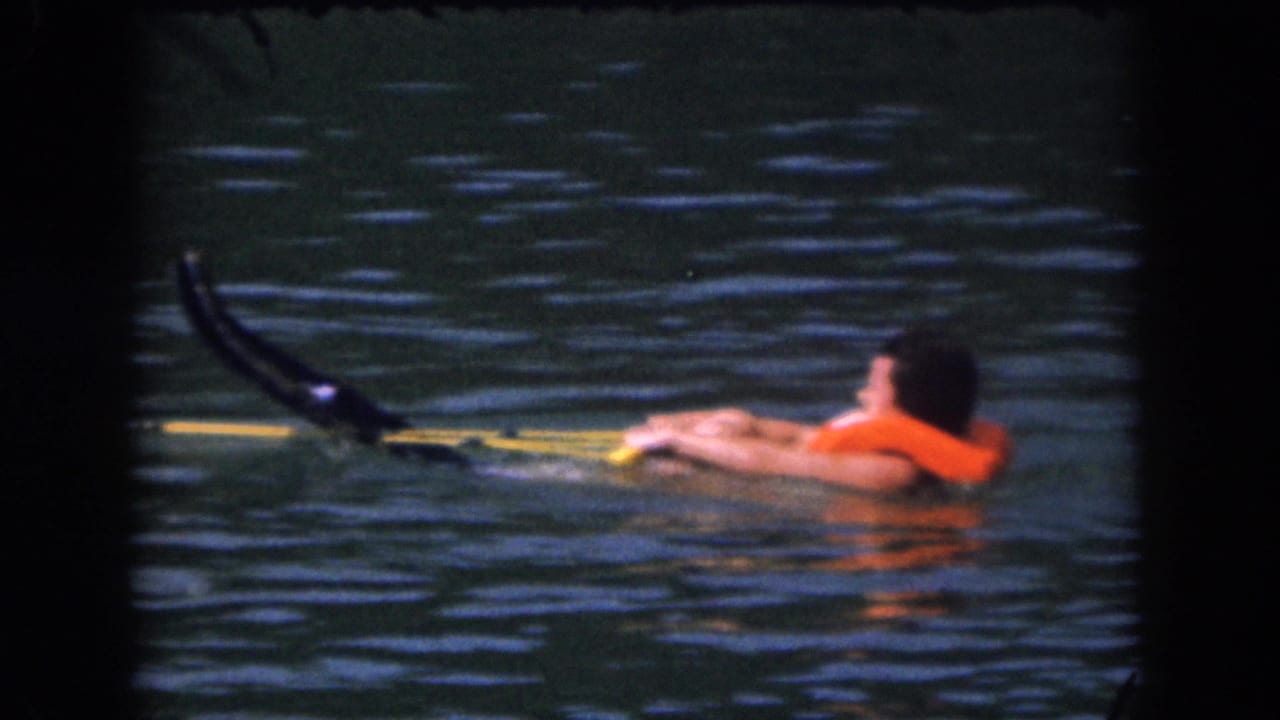 Person floating in lake water with orange life preserver, tow rope, and water skis partly visible.