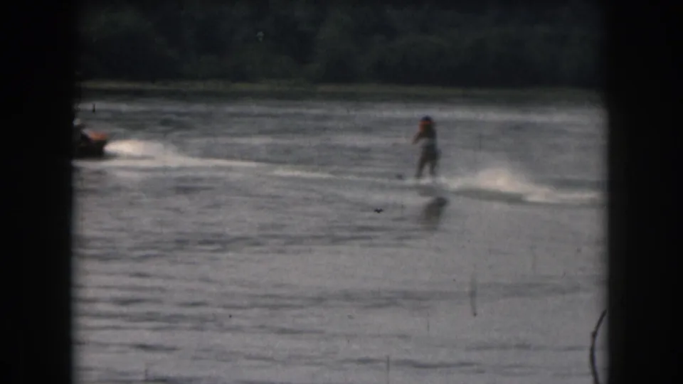 A distant skier is pulled behind a small motorboat on smooth water in this 1961 8mm Michigan lake recreation scene.