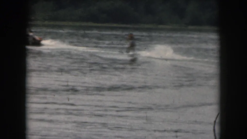 Soft 8mm frame showing a water skier behind a small boat, with motion blur and a tree-lined shoreline in the background.