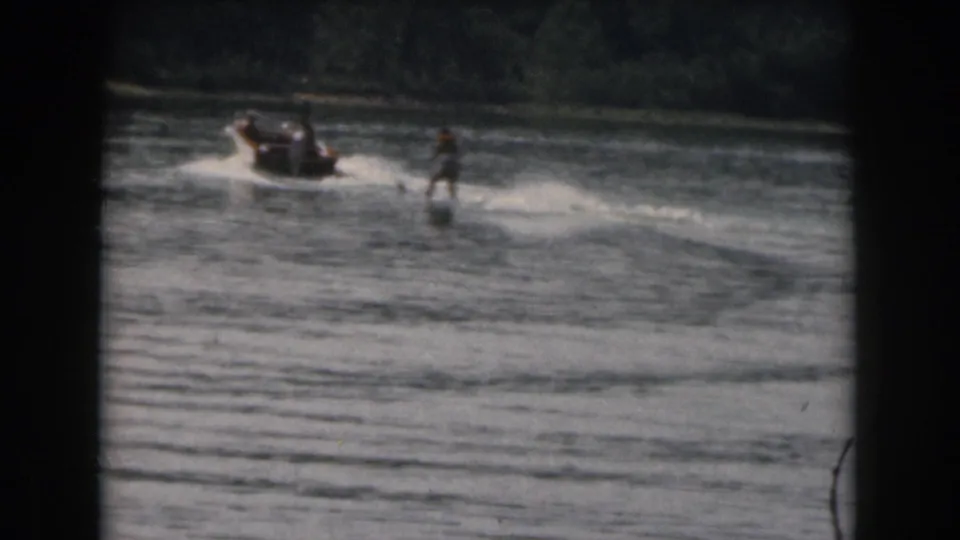 A small motorboat tows a distant water skier across a calm Michigan lake, with dark 8mm frame borders and muted archival color.