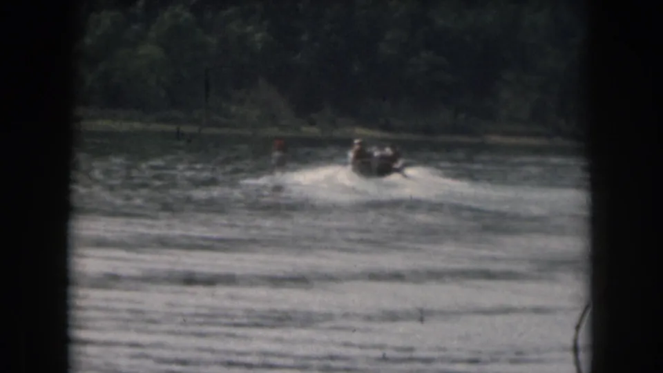 Distant 1961 Michigan home-movie frame of a boat towing a skier across rippled water beside a wooded shoreline.
