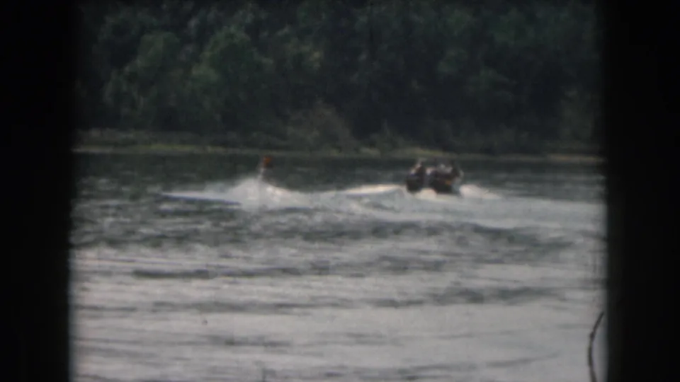 8mm home-movie frame from Grand Rapids, Michigan showing a water skier behind a small boat on a wooded lake.