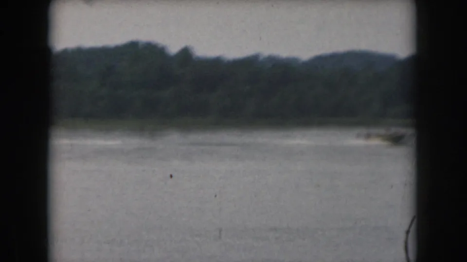 Wide 1961 Michigan lake frame with a distant small boat near the right edge and a wooded shoreline beyond.