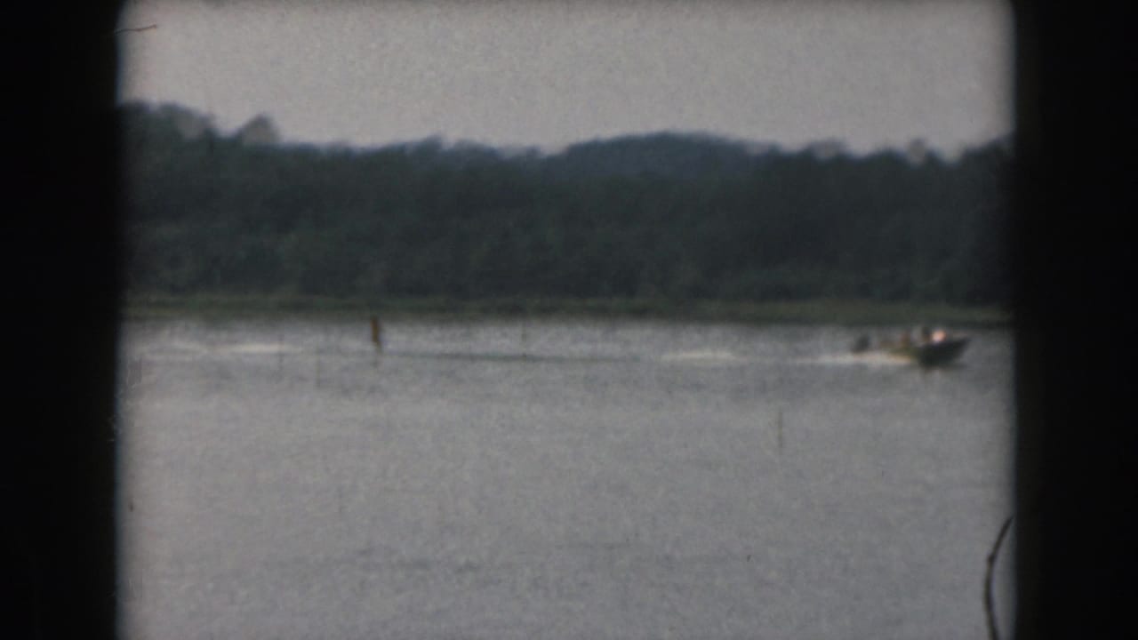 1961 Grand Rapids 8mm frame showing a distant water skier and motorboat on a lake below a tree-lined shore.