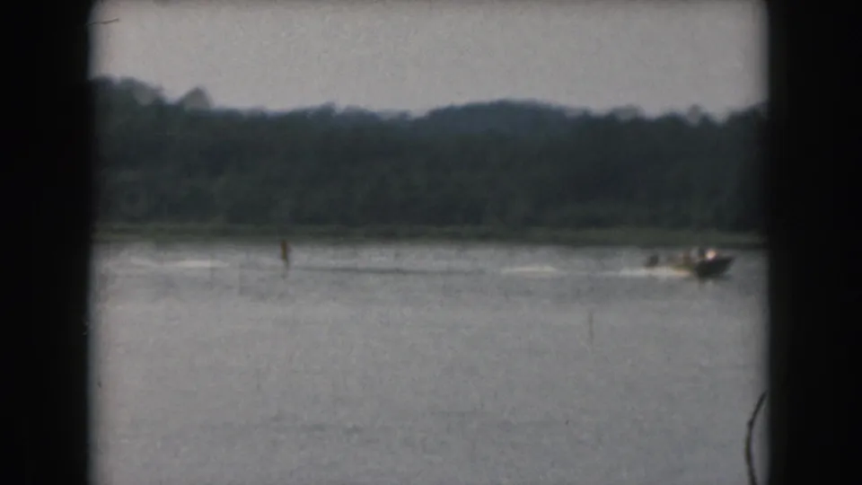 1961 Grand Rapids 8mm frame showing a distant water skier and motorboat on a lake below a tree-lined shore.