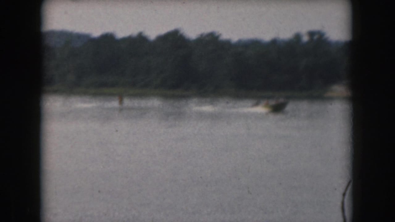 Motorboat speeds along the far side of a lake near the tree-lined shore.