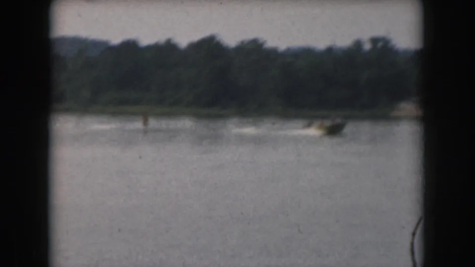 Distant motorboat and skier wake on a Grand Rapids lake, framed by dark 8mm film edges and a wooded horizon.