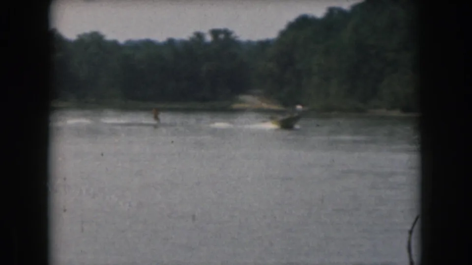 1961 home-movie frame of a water skier and nearby motorboat moving across a wooded Michigan lake.