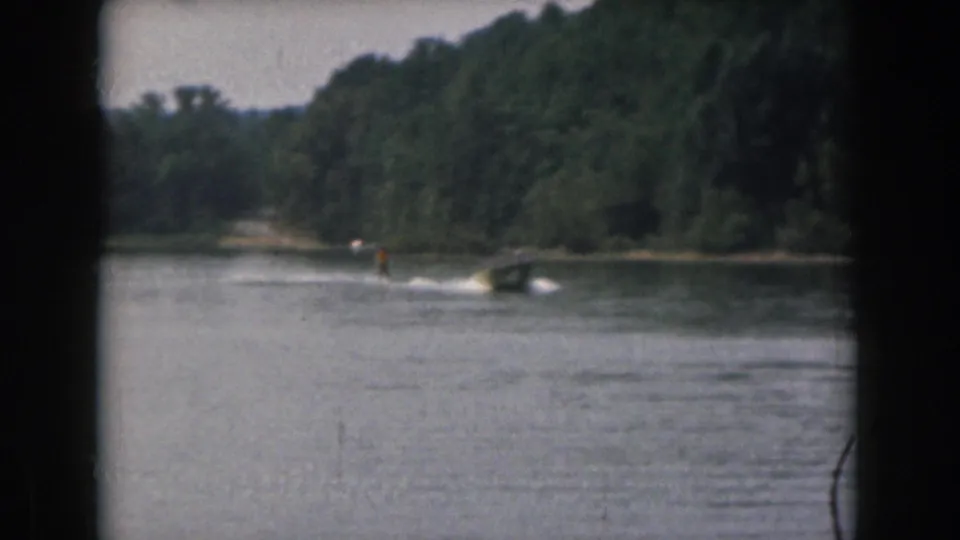 Distant water skier and motorboat on a Grand Rapids lake, with wooded shoreline and visible 8mm gate edges.