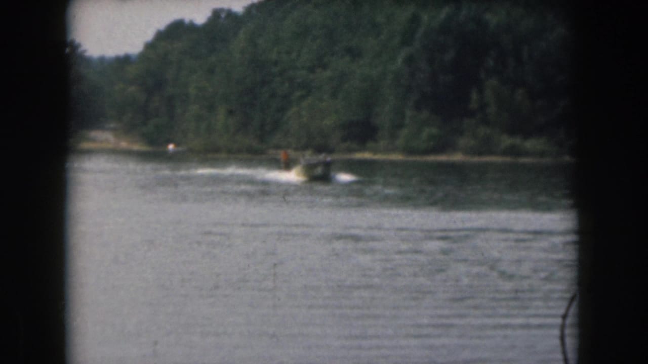 Small motorboat pulls a distant water skier across a calm lake near a wooded shoreline.