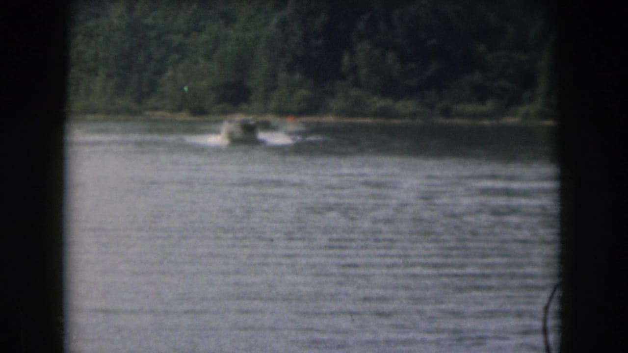 Motorboat travels across the lake, leaving a white wake near the wooded far shore.