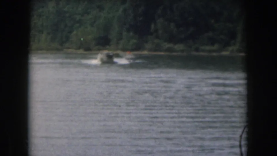 1961 Michigan lake scene with a small motorboat moving past a wooded shoreline and pulling a skier out of clear detail.