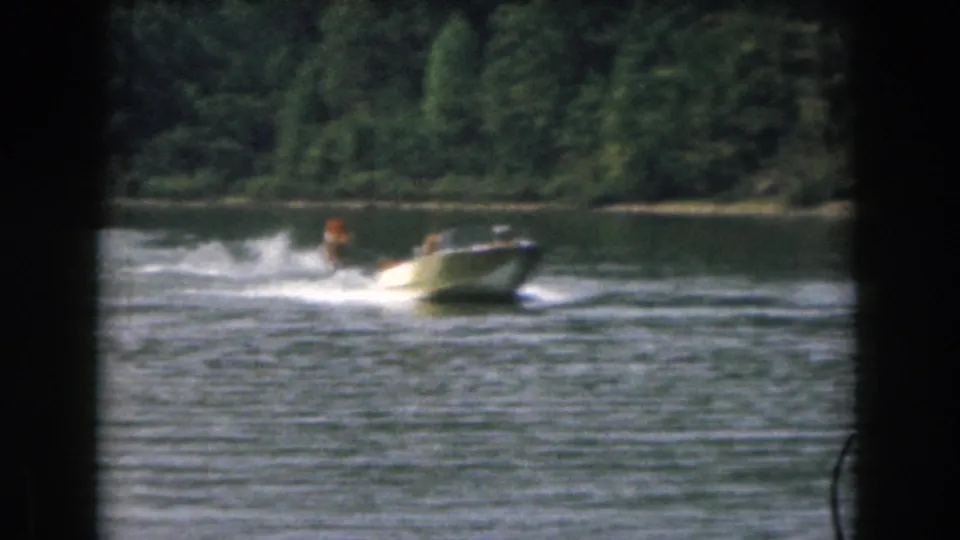 1961 Michigan lake scene with a small motorboat towing a skier in an orange vest past a wooded shoreline.