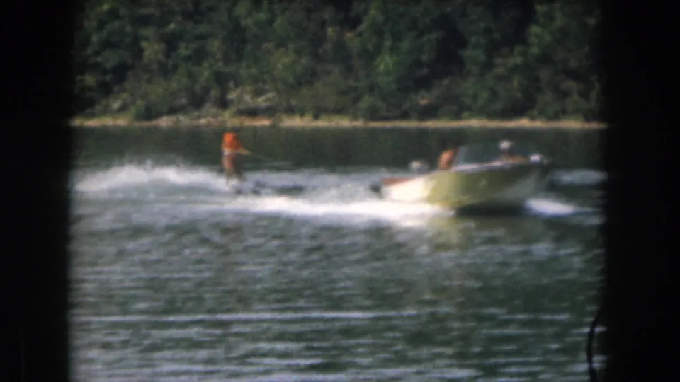 1961 Michigan home-movie frame of a boat towing a skier across a lake, with dark 8mm borders visible.