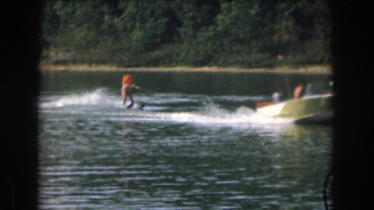Woman in an orange life vest water skis behind a small motorboat on a tree-lined lake.