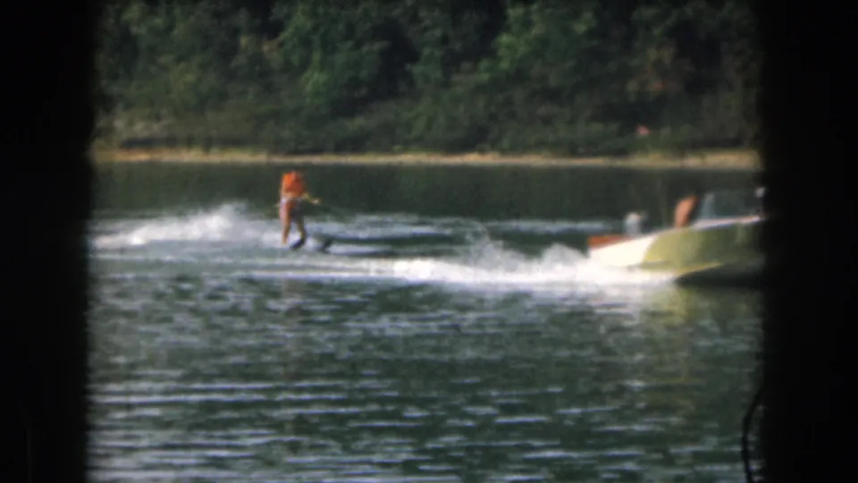 1961 home-movie frame from Grand Rapids, Michigan showing a woman water skiing behind a small motorboat across a wooded lake.