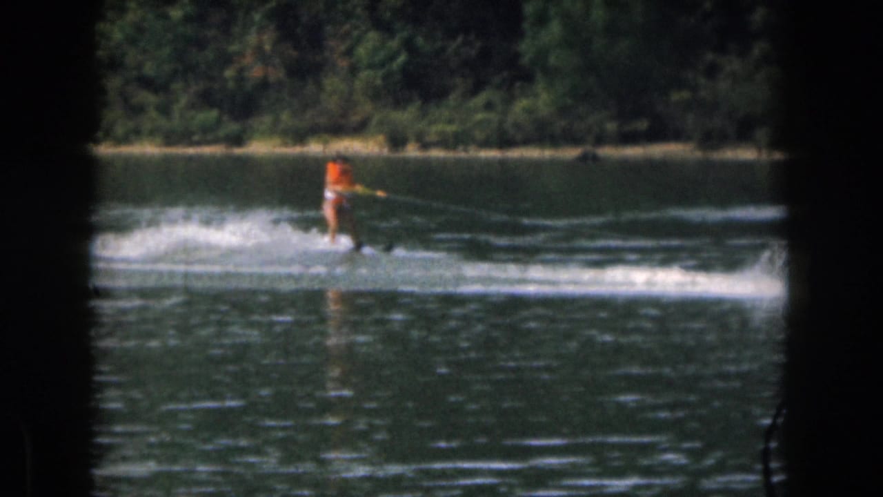 Water skier in an orange life vest glides across the lake with a tow rope extending off frame.