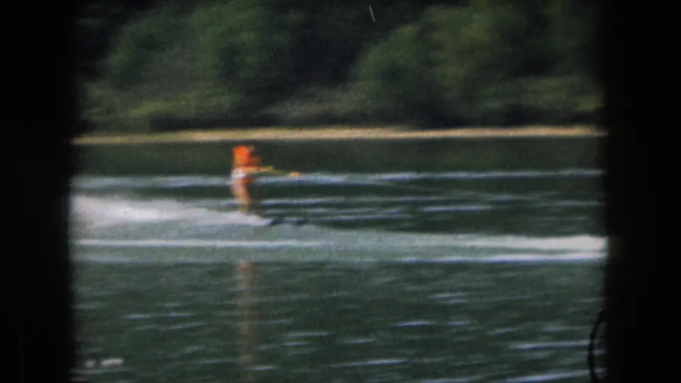 Soft 8mm frame of the woman water skiing across a Michigan lake, wearing an orange life vest and white swimwear.