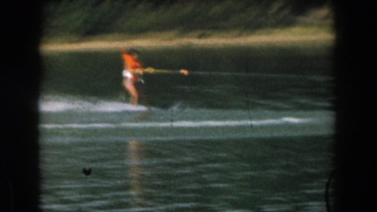 Distant water skier in an orange life vest moving across a calm lake.