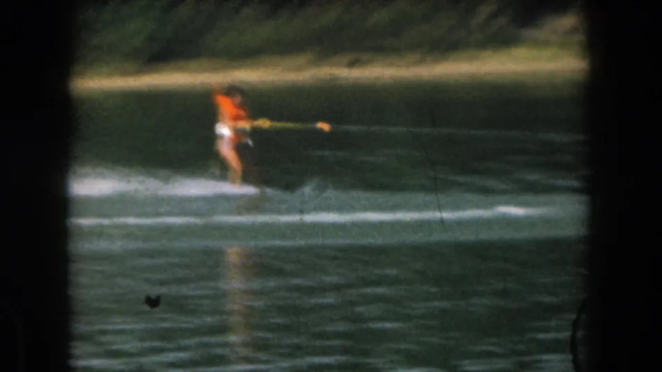 1961 home-movie frame from Grand Rapids, Michigan showing a distant water skier crossing a calm lake near a wooded shore.