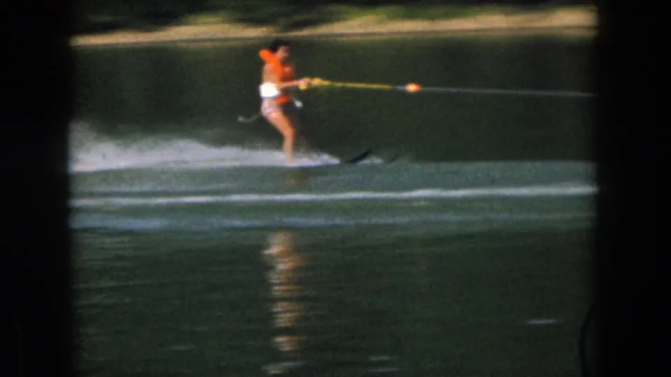 A distant skier in light swimwear and an orange life vest rides over the lake surface in this 1961 Michigan home-movie frame.