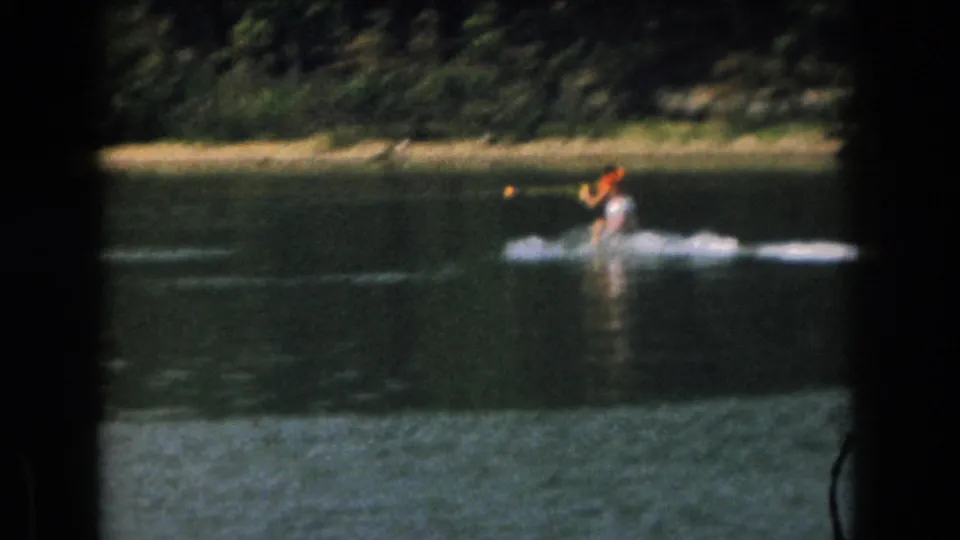 A 1961 8mm frame shows the skier low over the water, with a tow rope extending across a wooded Michigan lake scene.