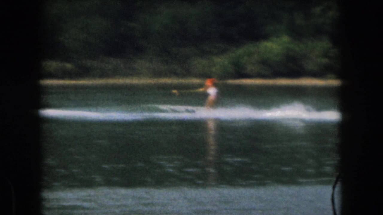 Distant water skier crossing a calm lake with a white wake and trees behind.