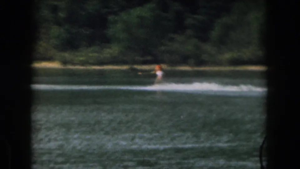 The 1961 home-movie frame shows a distant water skier continuing across the lake, with dark frame edges and soft 8mm grain.