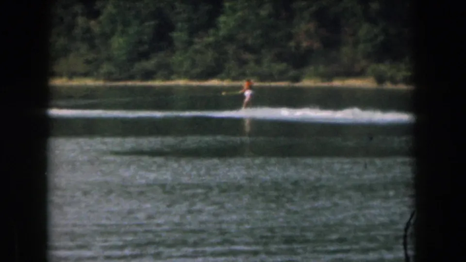 A distant water skier in the Grand Rapids lake footage rides across the frame, with a wooded shore and visible 8mm film texture.
