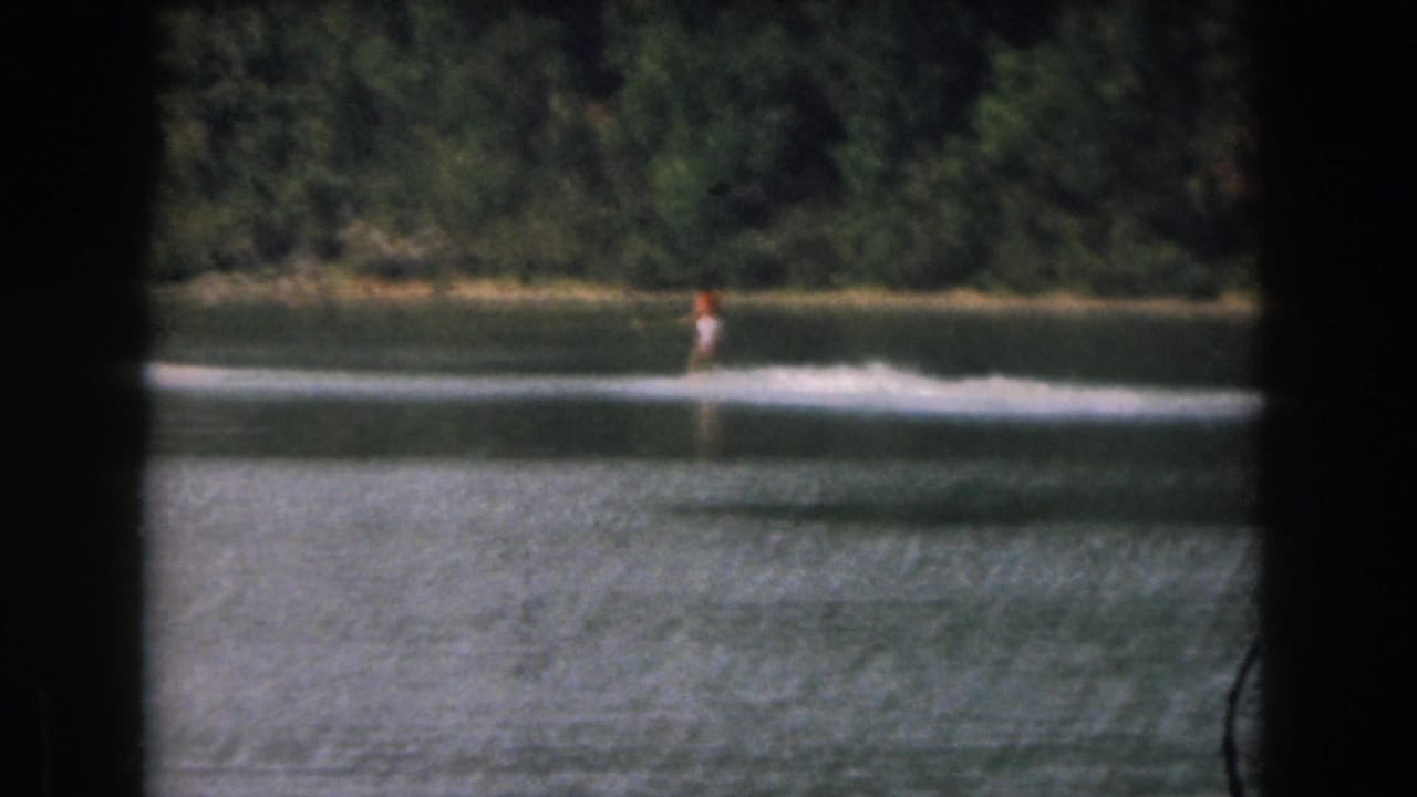1961 Grand Rapids-area home-movie frame showing a distant water skier moving across a calm wooded lake.