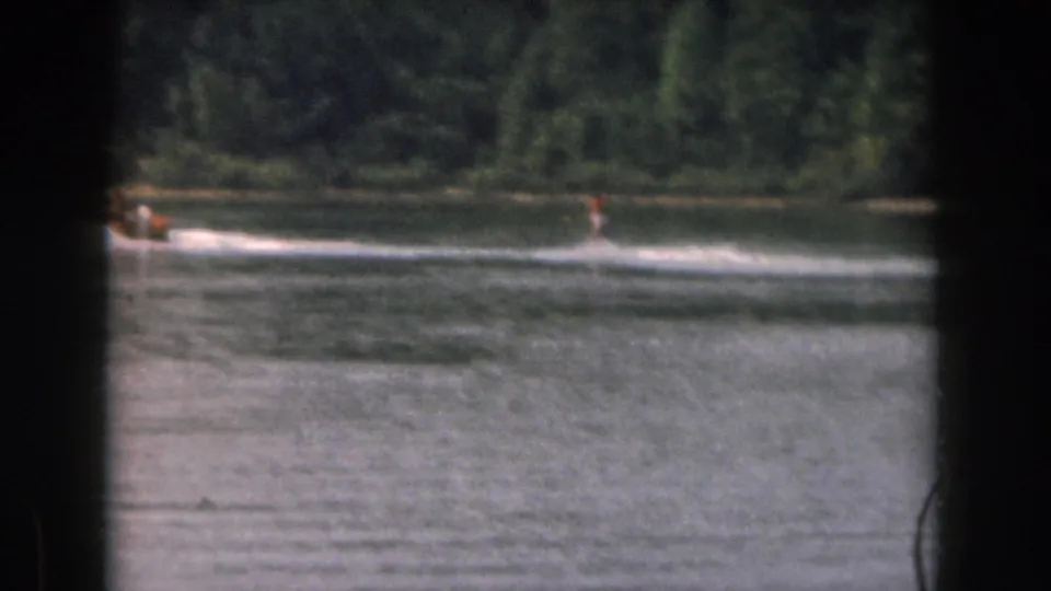 Home-movie frame from 1961 showing a small tow boat and distant water skier on a calm Michigan lake.