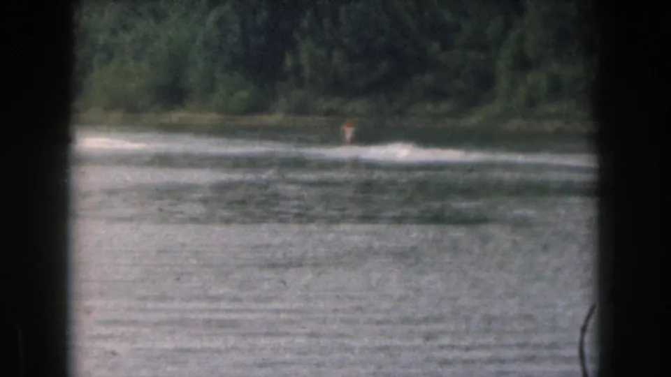 1961 Michigan home-movie still of a distant water skier passing a wooded shoreline on a calm lake.