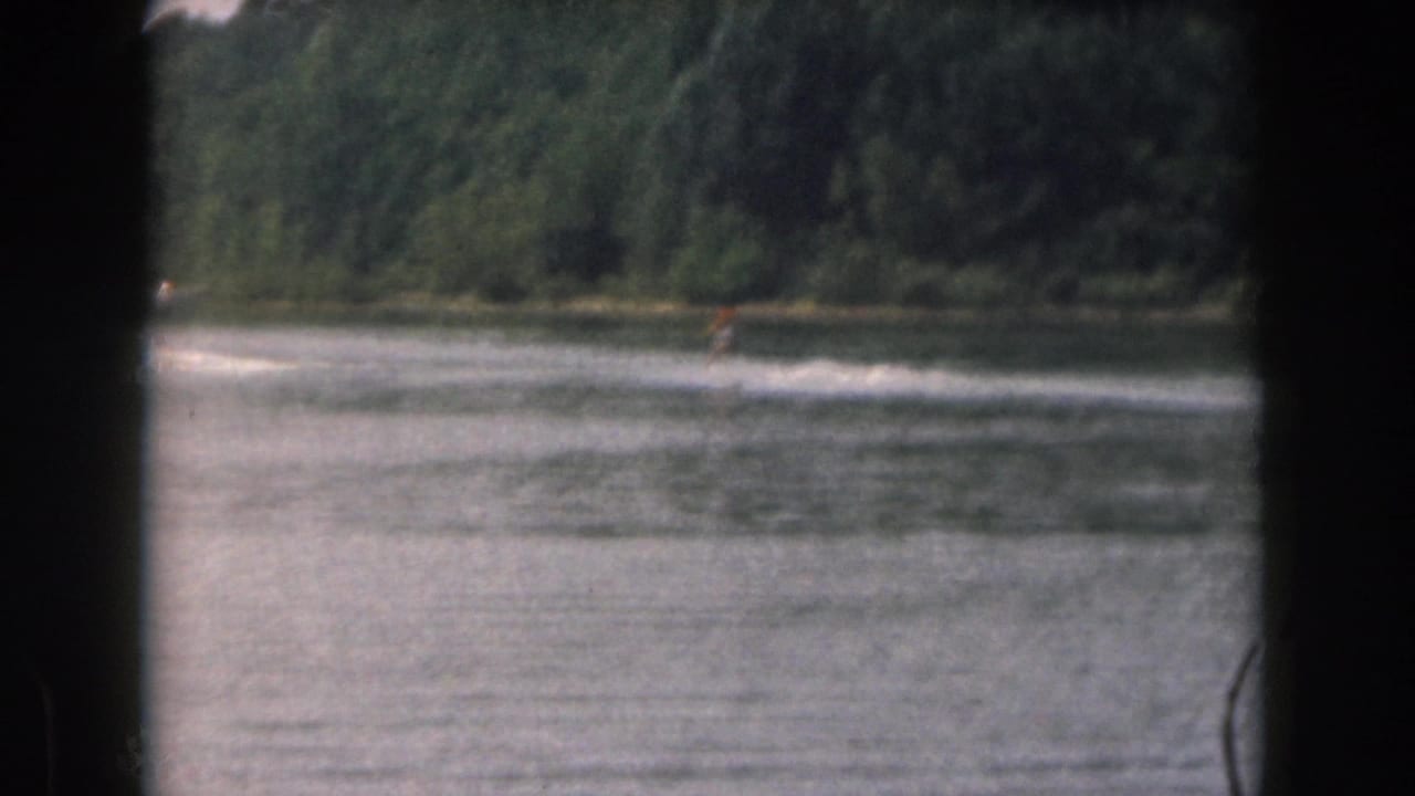 1961 Michigan home-movie frame showing a distant water skier moving across a lake with trees along the far shore.